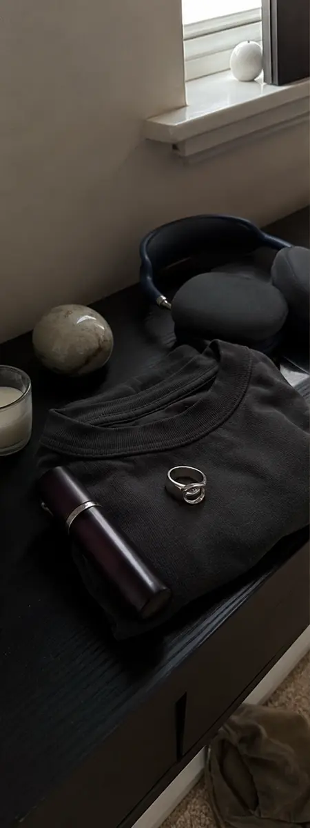 Phone-style dresser photo of a plum atomiser, folded charcoal T-shirt, rings and headphones